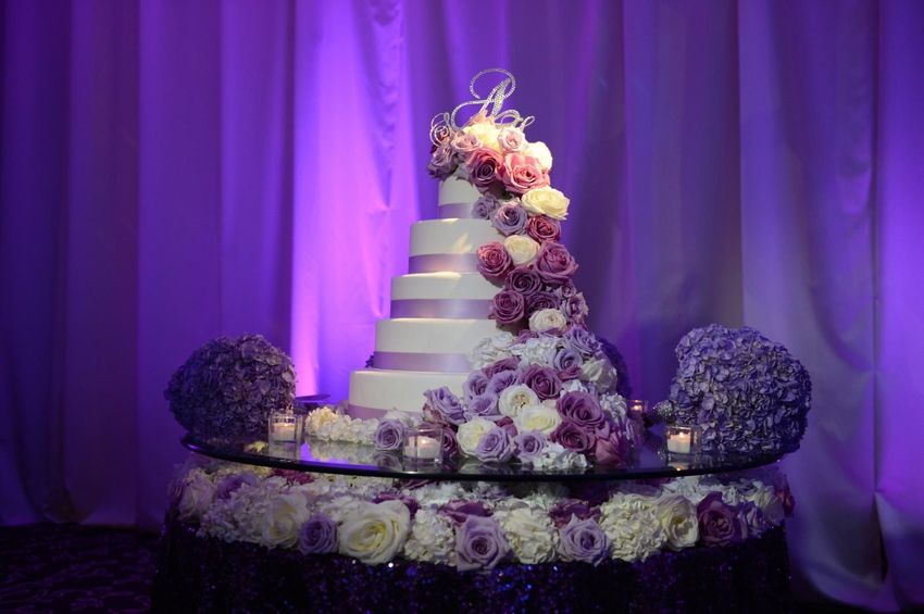 6- layered wedding cake adorned with white and purple roses on round glass cake stand decorated with purple hydrangea and votive candles